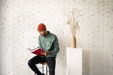 Man in knit hat reading book by dried plant vase against white brick wall - RCPF00507