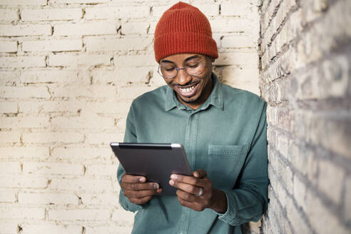 Glücklicher Mann, der ein digitales Tablet benutzt, während er sich an eine Ziegelmauer lehnt - RCPF00520