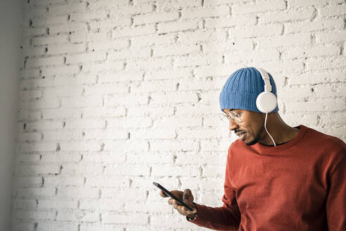 Lächelnder Mann, der ein Mobiltelefon benutzt und über Kopfhörer Musik hört, vor einer weißen Backsteinmauer - RCPF00529