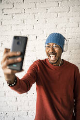 Cheerful man with mouth open taking selfie while listening music against white brick wall - RCPF00530