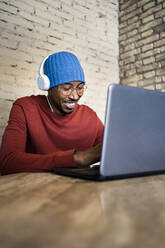 Lächelnde männliche Fachkraft, die einen Laptop benutzt und über Kopfhörer auf einem Tisch Musik hört - RCPF00535