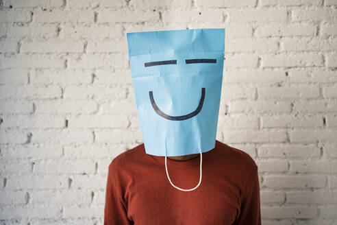 Mittlerer erwachsener Mann mit blauer Papiertüte im Gesicht an einer weißen Backsteinmauer - RCPF00538