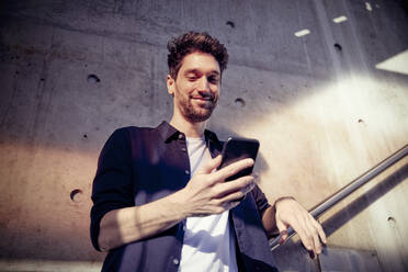 Smiling businessman using mobile phone while standing against wall - RHF02554
