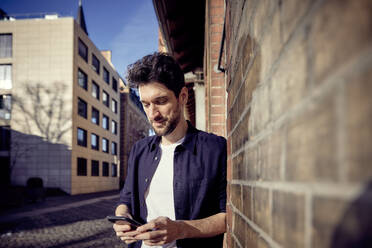 Businessman using mobile phone while leaning on wall in city - RHF02557