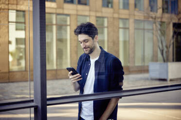 Smiling businessman using mobile phone while leaning on glass wall - RHF02561