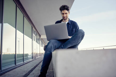 Businessman using laptop while sitting on retaining wall - RHF02569