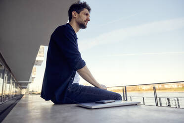 Geschäftsmann mit Blick auf die Aussicht, während er mit Laptop und Mobiltelefon auf einer Stützmauer sitzt - RHF02570