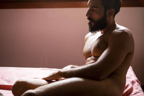 Young naked man sitting on bed at home - MIMFF00337