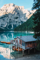 Majestic scenery of wooden house located on shore of clear lake with turquoise water and boats floating on smooth surface on background of mountains in morning light - ADSF19357