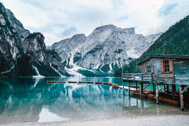 Majestic scenery of wooden house located on shore of clear lake with turquoise water and boats floating on smooth surface on background of mountains in morning light - ADSF19362