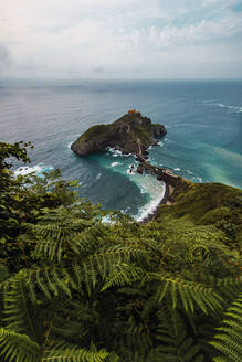 Drohnenansicht eines gepflasterten Weges, der über eine Steinbrücke und den Kamm eines felsigen Hügels zu einem einsamen Haus auf der Insel Gaztelugatxe führt, umgeben von ruhigem Meerwasser unter bewölktem Himmel im Baskenland - ADSF19379