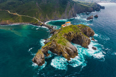 Drohnenansicht eines gepflasterten Weges, der über eine Steinbrücke und den Kamm eines felsigen Hügels zu einem einsamen Haus auf der Insel Gaztelugatxe führt, umgeben von ruhigem Meerwasser unter bewölktem Himmel im Baskenland - ADSF19381