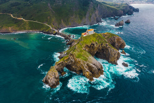 Drohnenansicht eines gepflasterten Weges, der über eine Steinbrücke und den Kamm eines felsigen Hügels zu einem einsamen Haus auf der Insel Gaztelugatxe führt, umgeben von ruhigem Meerwasser unter bewölktem Himmel im Baskenland - ADSF19381