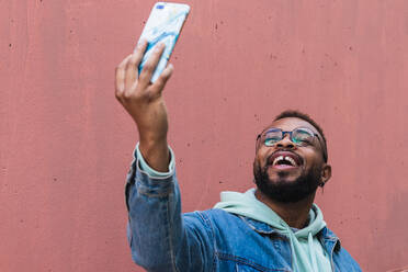Low angle cheerful adult black hipster guy in casual outfit taking self portrait on mobile phone against red background - ADSF19393
