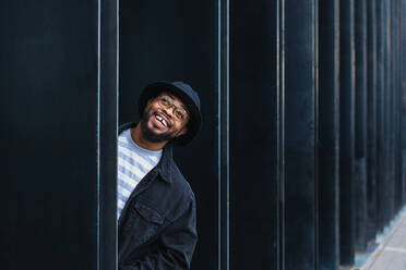 Optimistic adult black hipster male in hat and glasses peeking out of pillar of modern urban building and looking away with curiosity - ADSF19396