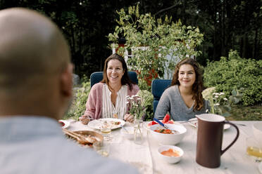 Smiling woman and daughter looking at father while sitting by dining table in yard - MASF20996