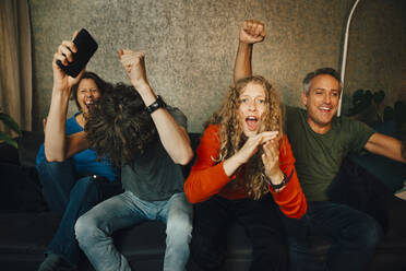 Happy parents with children cheering while watching sports in living room at night - MASF21002