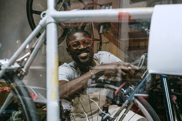 Porträt eines lächelnden Besitzers, der ein Fahrrad in einer Werkstatt repariert, gesehen durch ein Glasfenster - MASF21061