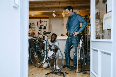 Lächelnder Besitzer, der in der Werkstatt ein Fahrrad repariert, gesehen durch ein Glasfenster - MASF21063