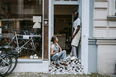 Senior owner talking to colleague while sitting on steps of bicycle shop - MASF21067