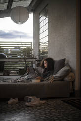 Seitenansicht eines weiblichen Teenagers, der ein digitales Tablet benutzt, während er sich auf dem Sofa zu Hause entspannt - MASF21080