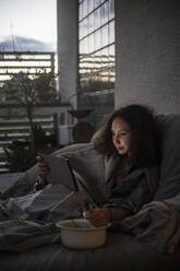 Weiblicher Teenager, der ein digitales Tablet benutzt, während er zu Hause auf dem Sofa isst - MASF21081