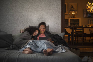 Mother and daughter watching digital tablet together while relaxing on sofa at home - MASF21083