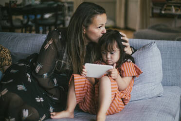 Mutter küsst Tochter mit Down-Syndrom, die auf dem Sofa zu Hause ihr Smartphone benutzt - MASF21088