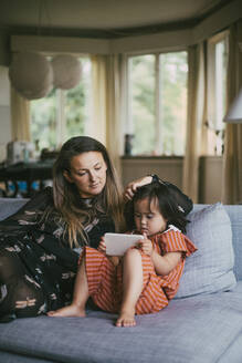 Mutter schaut auf ihre Tochter mit Down-Syndrom, die ein Mobiltelefon benutzt, während sie auf dem Sofa im Wohnzimmer sitzt - MASF21089