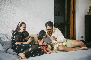 Father and disabled daughter using digital tablet while smiling girl embracing mother on sofa at home - MASF21103