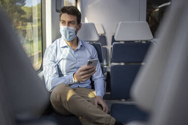 Geschäftsmann mit Gesichtsmaske, der wegschaut, während er im Zug sitzend ein Mobiltelefon benutzt - IFRF00216