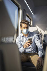 Businessman with protective face mask using mobile phone while sitting in train during COVID-19 - IFRF00217