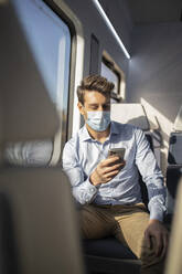 Geschäftsmann mit Schutzmaske, der im Zug sitzend ein Mobiltelefon benutzt, während COVID-19 - IFRF00217
