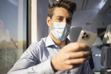 Mid adult businessman with face mask using smart phone while traveling by train during COVID-19 - IFRF00218