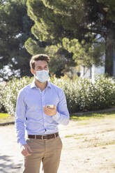 Businessman wearing protective face mask using mobile phone while standing on footpath - IFRF00222
