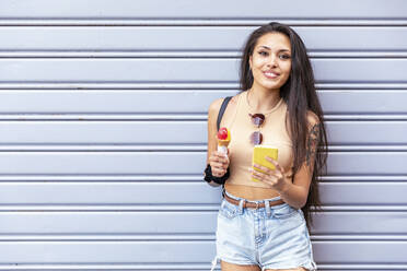 Smiling woman with ice cream cone using mobile phone while standing against shutter - WPEF03826