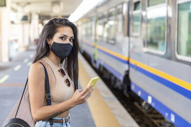 Young woman wearing face mask using mobile phone while standing on platform - WPEF03829