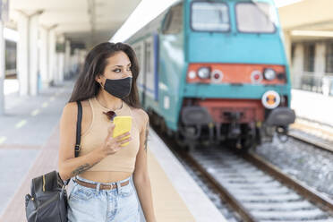 Young woman wearing face mask looking away while using mobile phone standing at station - WPEF03831