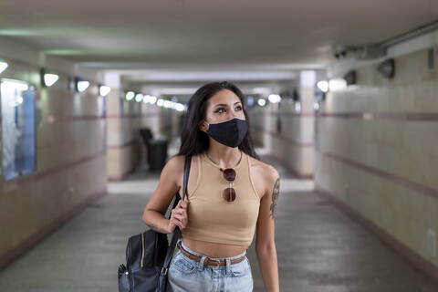 Junge Frau mit Gesichtsmaske, die eine Tasche trägt, während sie in einer unterirdischen Bahnhofspassage geht, lizenzfreies Stockfoto