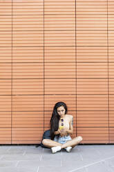 Junge Frau benutzt ein Mobiltelefon, während sie an der Wand sitzt - WPEF03845