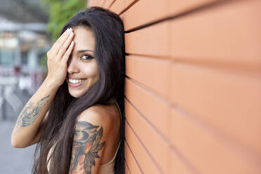 Young woman smiling while covering face with hand leaning on wall - WPEF03850