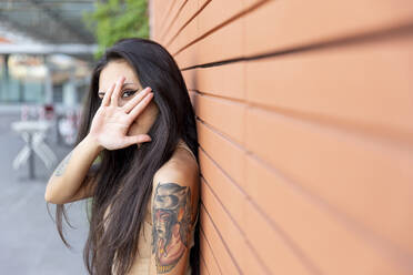 Woman looking through finger while leaning on wall - WPEF03851