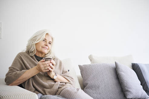 Nachdenkliche Frau im Ruhestand hält eine Kaffeetasse, während sie auf dem Sofa vor einer weißen Wand in einer Wohnung sitzt - JAHF00032