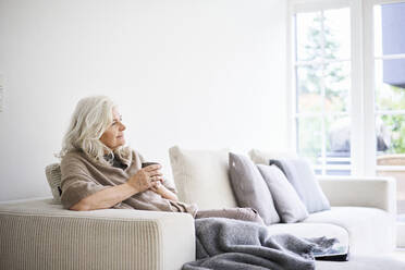 Nachdenkliche Frau mit langen weißen Haaren hält eine Kaffeetasse, während sie auf dem Sofa in einer Wohnung sitzt - JAHF00033