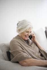 Cheerful senior woman talking on mobile phone while sitting against white wall at home - JAHF00034