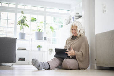 Ältere Frau im Ruhestand sitzt auf dem Boden und benutzt ein digitales Tablet im Wohnzimmer einer Wohnung - JAHF00038