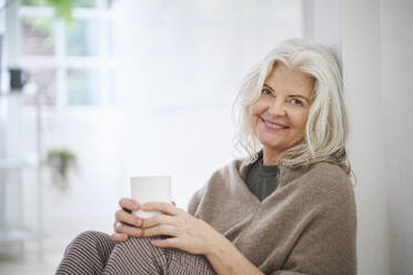 Glückliche ältere Frau hält eine Kaffeetasse, während sie zu Hause im Wohnzimmer sitzt - JAHF00041