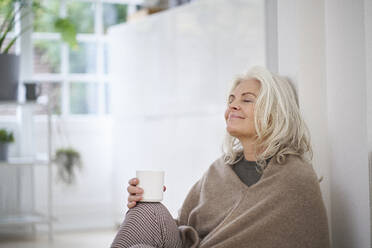 Ältere Frau im Ruhestand hält eine Kaffeetasse, während sie mit geschlossenen Augen zu Hause sitzt und träumt - JAHF00042