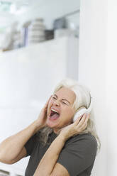 Cheerful woman enjoying music through headphones while shouting at apartment - JAHF00044