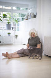 Frau im Ruhestand sitzt auf dem Boden und betrachtet die Katze in der Wohnung - JAHF00045